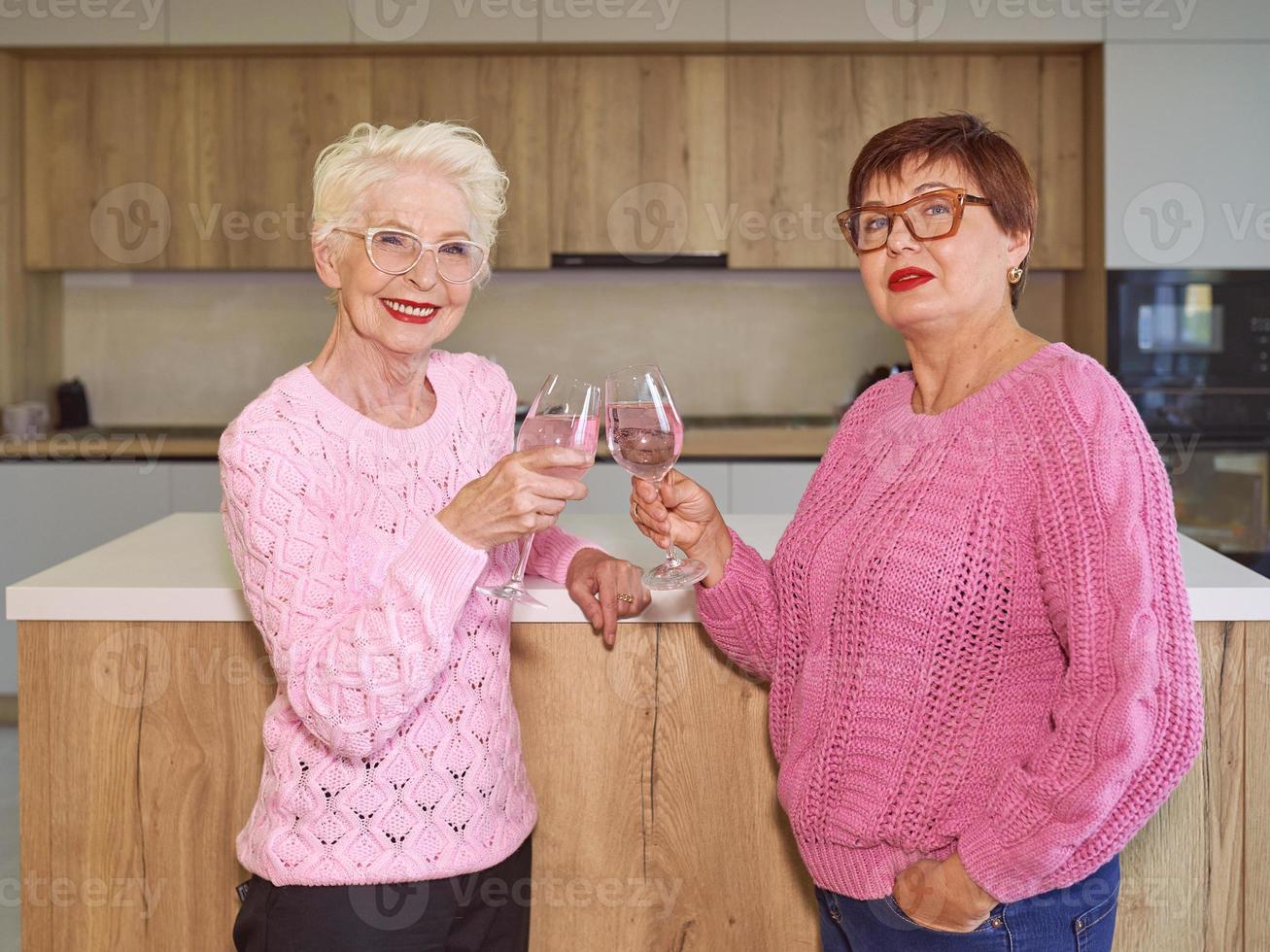 Dos mujeres mayores con estilo en suéteres de color rosa bebiendo vino rosado en la cocina moderna chismeando. amistad, hablar, chismes, eventos, relaciones, noticias, concepto de familia foto