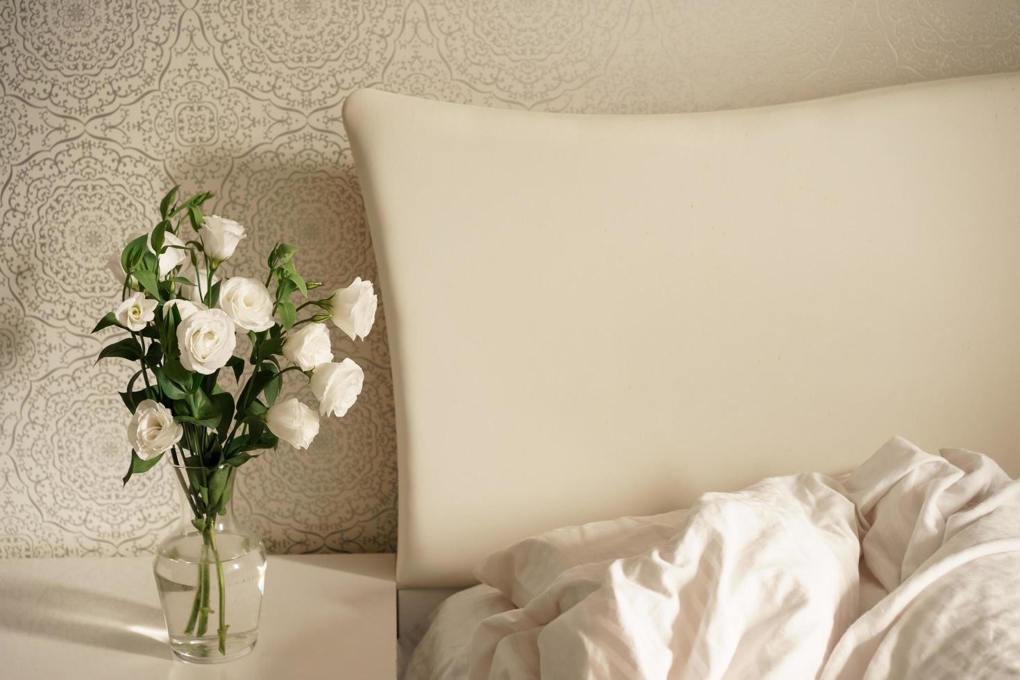 flores blancas en un jarrón de vidrio transparente se colocan en una mesita de noche junto a una cama con una gran cabecera blanca. interior del dormitorio. habitacion moderna foto