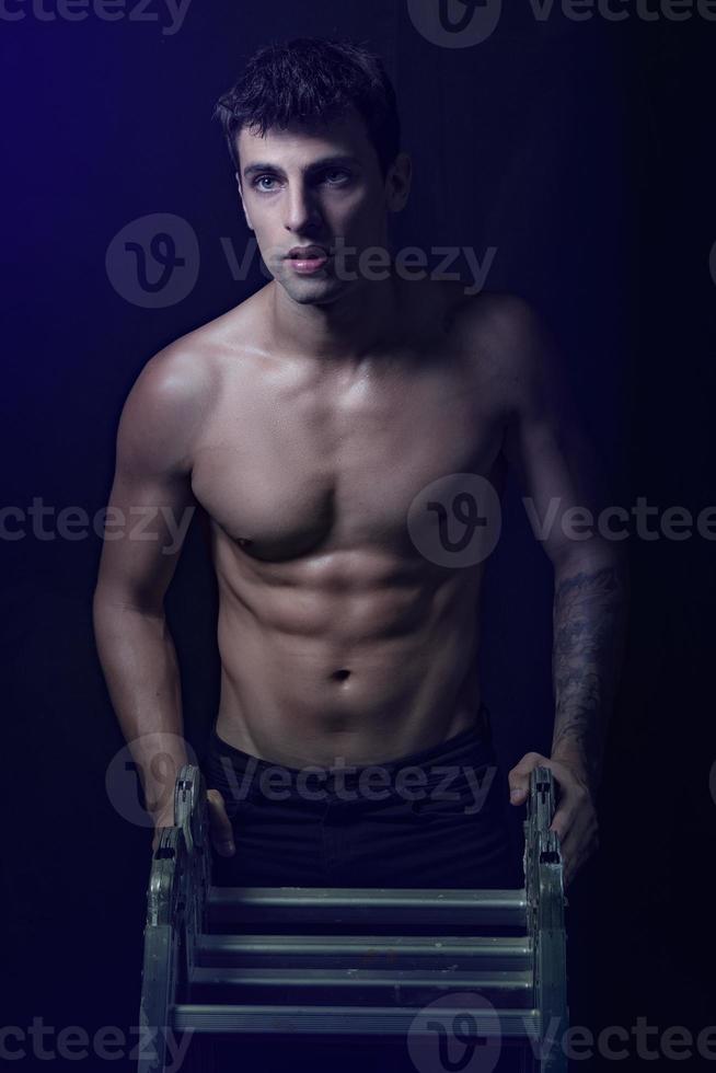 Blue eyed handsome man standing leaning on metal ladder, looking to the side, naked torso. photo