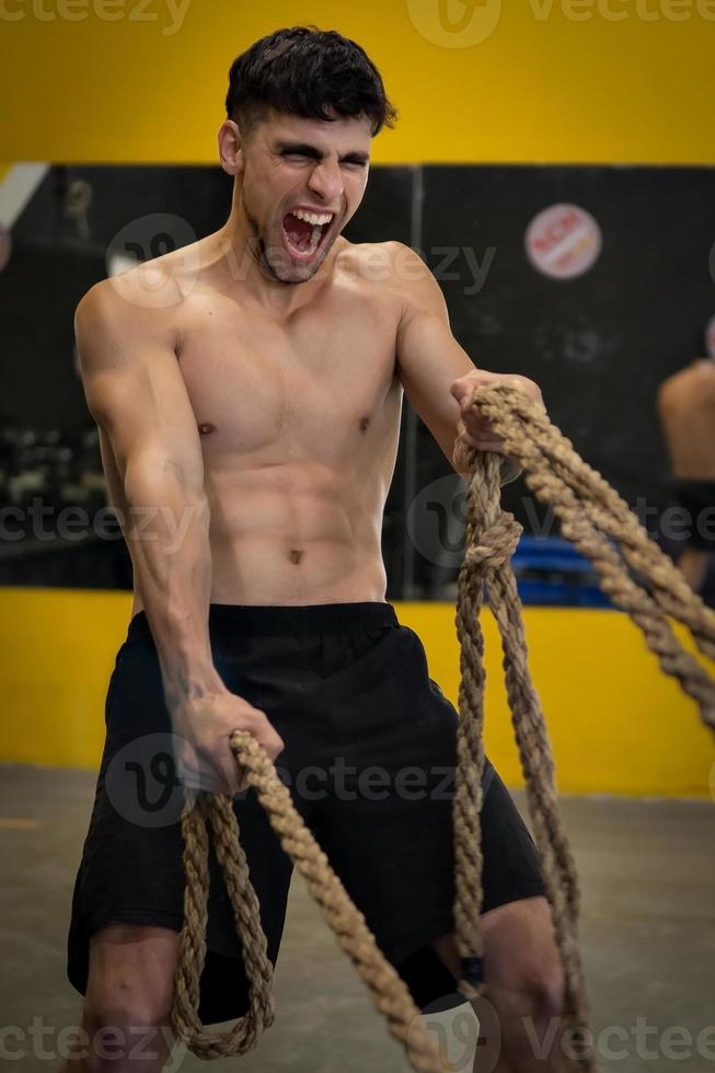 Hombre agresivo poderoso muscular que trabaja con una cuerda en el gimnasio de entrenamiento funcional foto