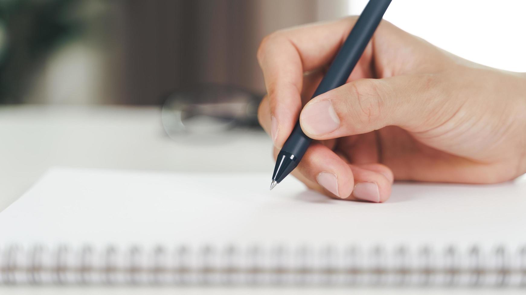 Cerca de joven en manos de tela casual escribiendo en el bloc de notas, cuaderno con bolígrafo sobre la mesa. foto