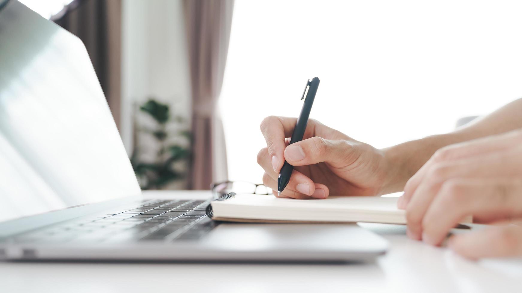 Close up of young man in casual cloth hands writing down on the notepad, notebook using ballpoint pen with laptop computer on the table. photo