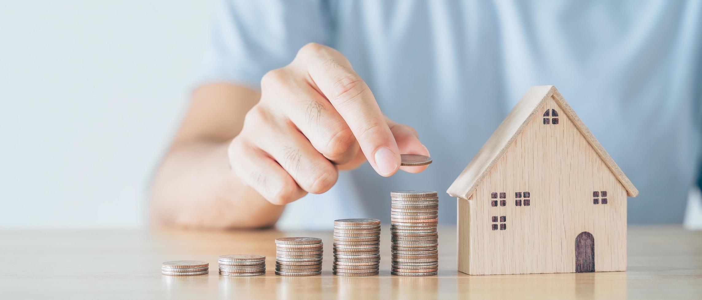 La mano del hombre puso la moneda en la pila de monedas con la casa de madera en la mesa de madera. Ahorrar dinero para comprar casa, concepto de préstamo hipotecario de plan financiero. foto