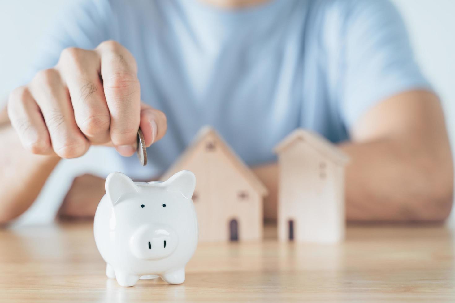 La mano del hombre puso una moneda en una alcancía blanca con una casa de madera en la mesa de madera. Ahorrar dinero para comprar casa, concepto de préstamo hipotecario de plan financiero. foto