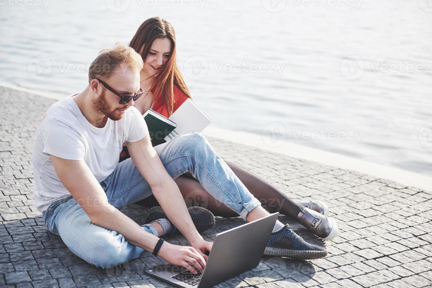 Dos estudiantes, chico y chica, están sentados al aire libre y disfrutando de una computadora portátil, estudiando al aire libre en un día soleado foto