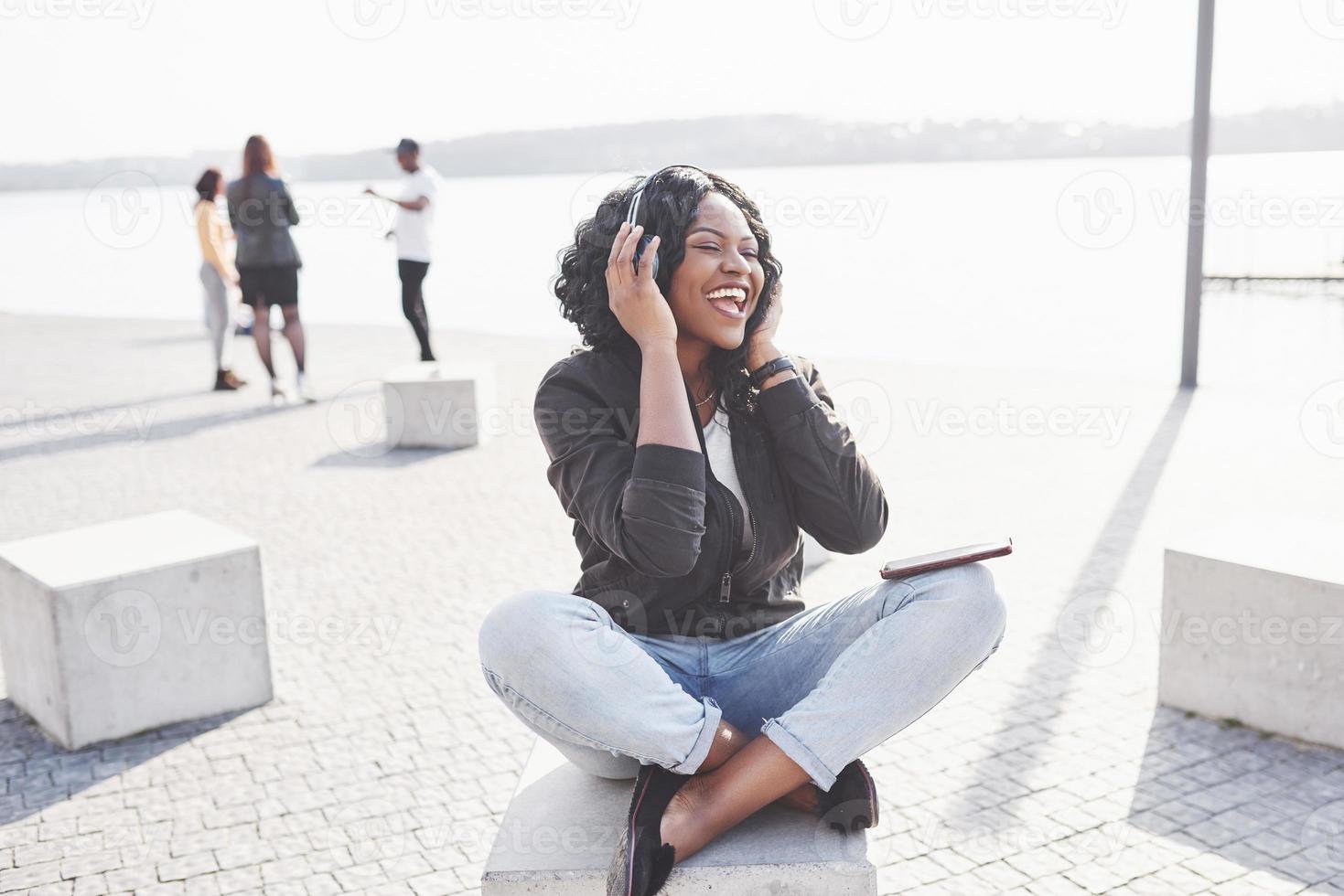 Portrait of a beautiful young pretty African American girl sitting on the beach or lake and listening to music in her headphones photo