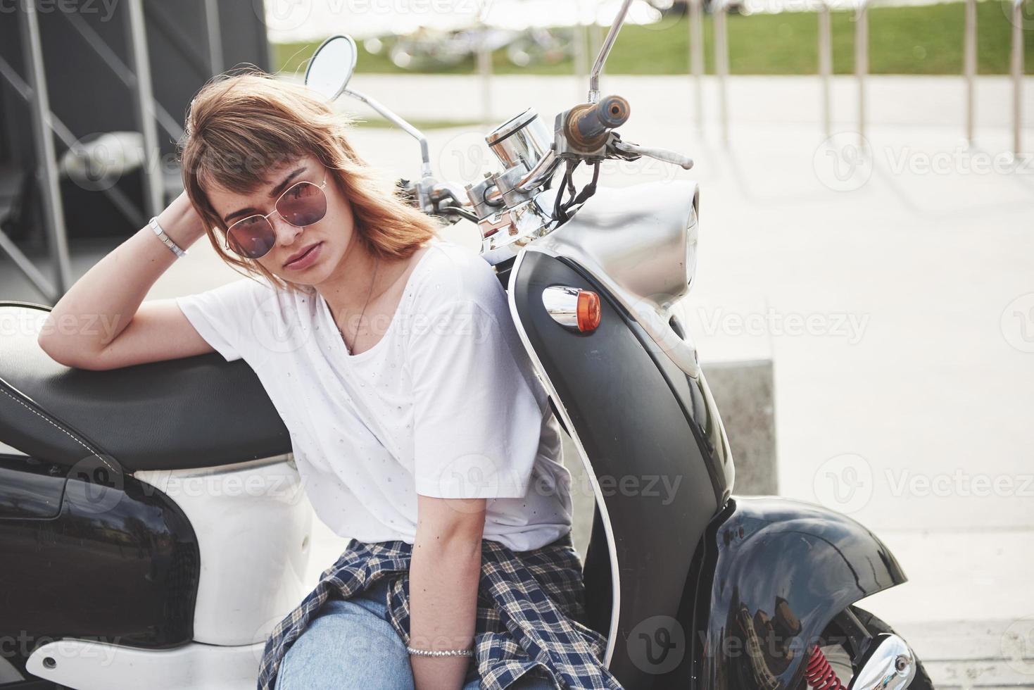 Retrato de una hermosa chica hipster sentada en un scooter retro negro, sonriendo posando y disfrutando del cálido sol primaveral. foto