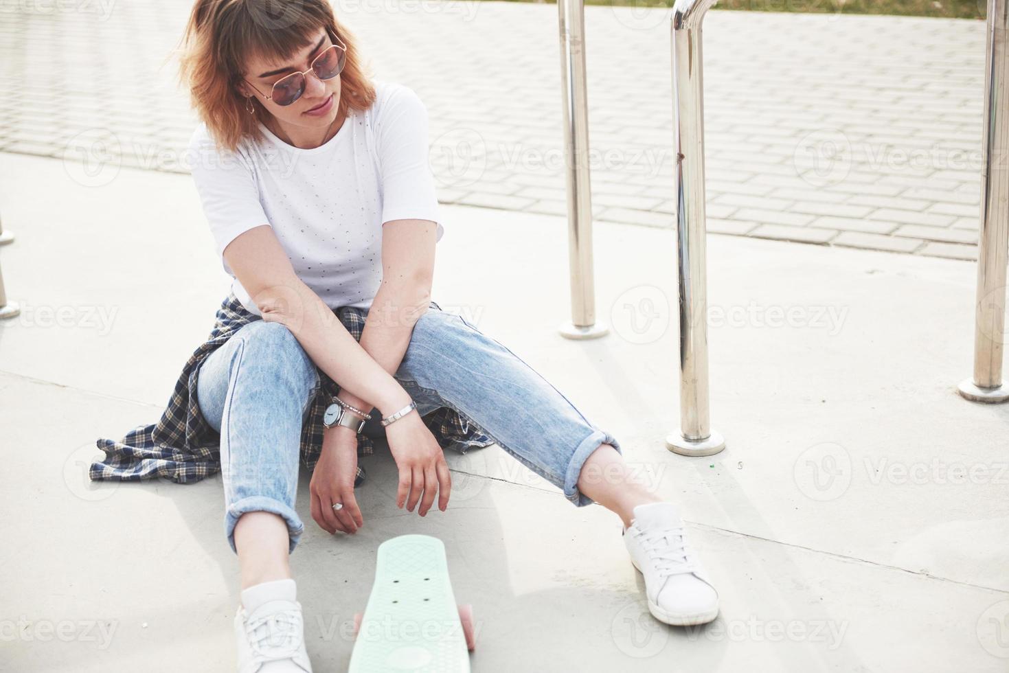 una foto de una hermosa chica con un cabello hermoso sostiene una patineta sobre una tabla larga, mirando a la cámara y sonriendo, la vida urbana.