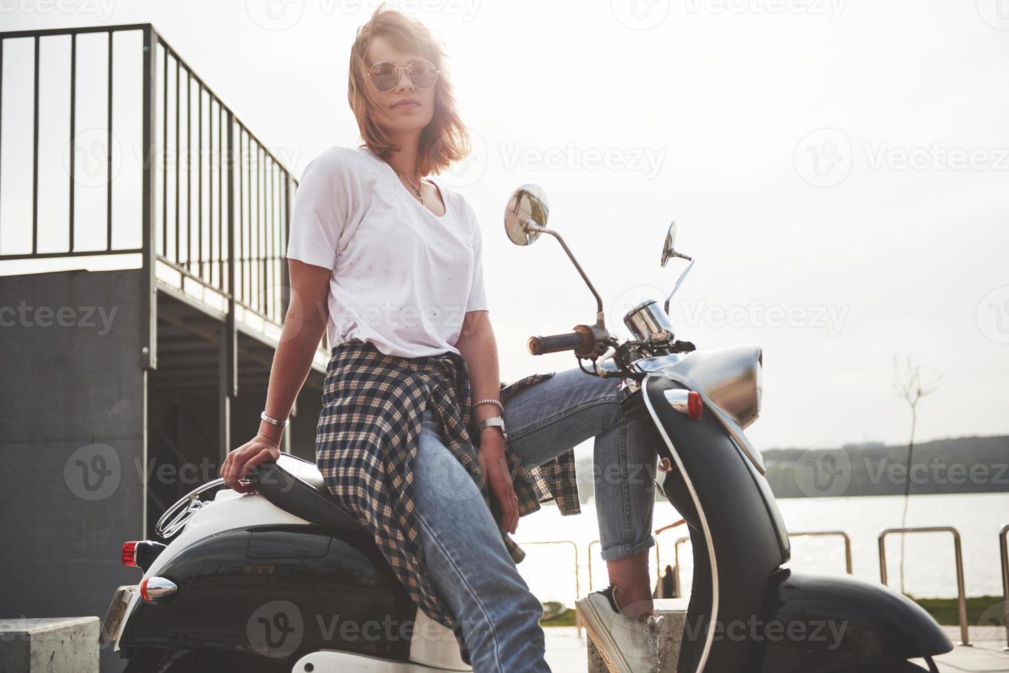 Portrait of a beautiful girl hipster sitting on a black retro scooter, smiling posing and enjoy the warm spring sunshine. photo