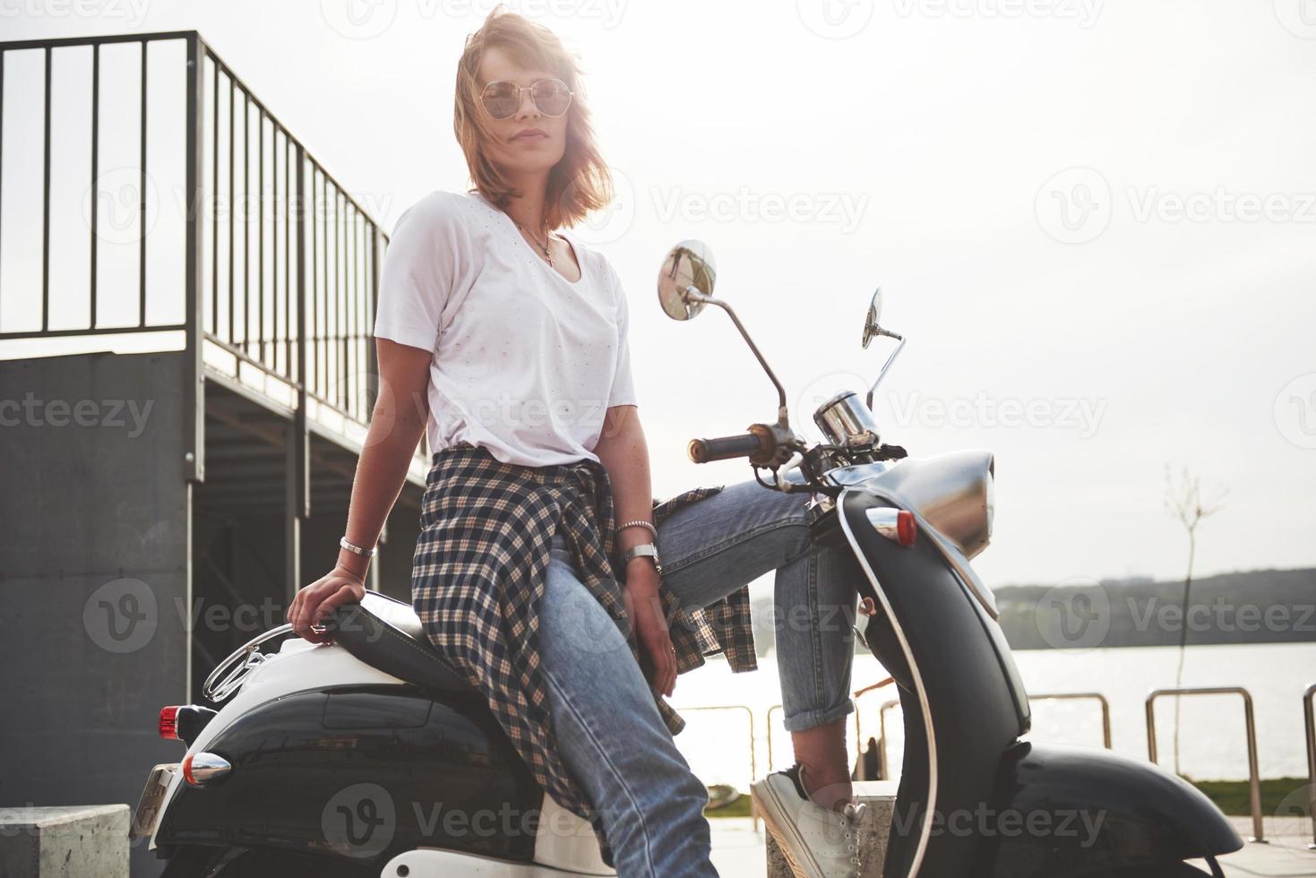 Retrato de una hermosa chica hipster sentada en un scooter retro negro, sonriendo posando y disfrutando del cálido sol primaveral. foto