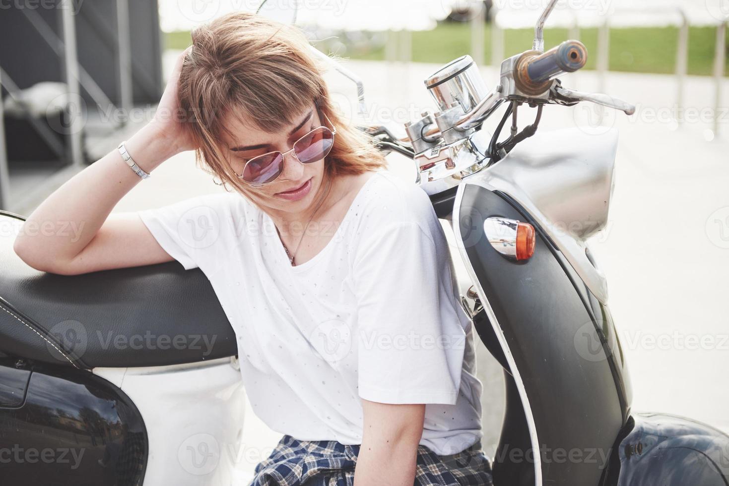 Retrato de una hermosa chica hipster sentada en un scooter retro negro, sonriendo posando y disfrutando del cálido sol primaveral. foto