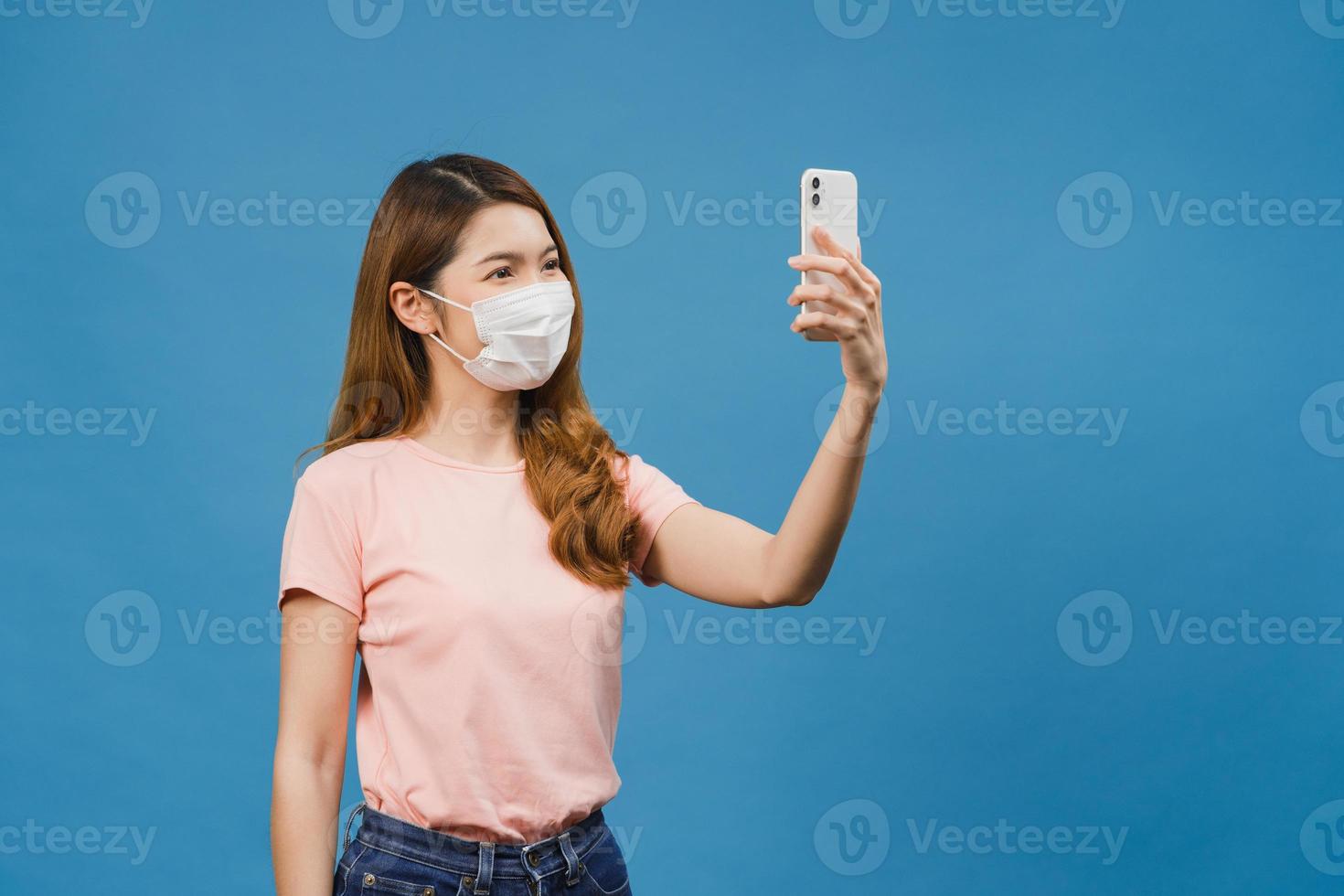 Sonriente mujer asiática adorable con mascarilla médica haciendo foto selfie en teléfono inteligente con expresión positiva en ropa casual y soporte aislado sobre fondo azul.
