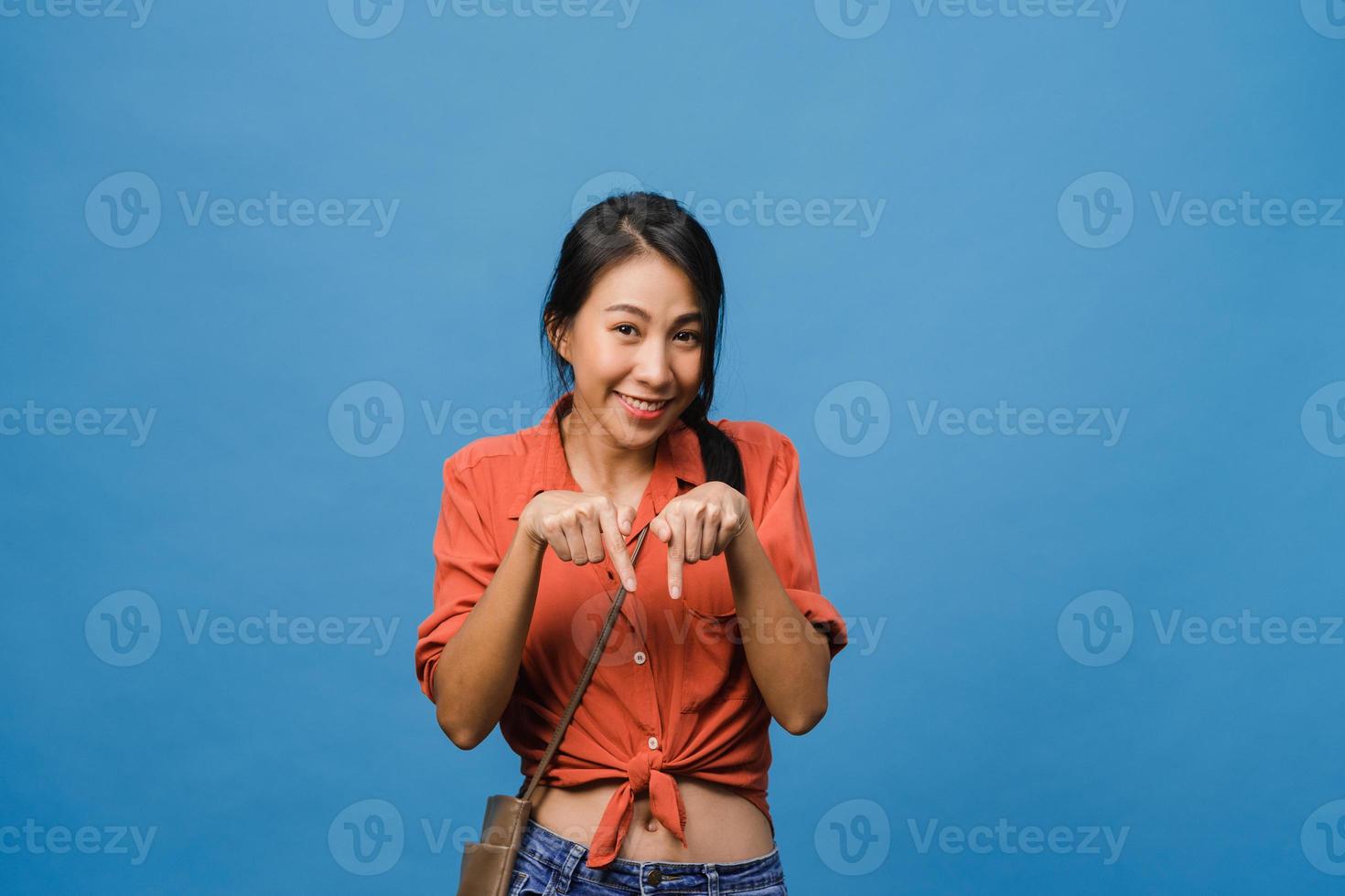 Portrait of young Asian lady smiling with cheerful expression, shows something amazing at blank space in casual cloth and looking at camera isolated over blue background. Facial expression concept. photo