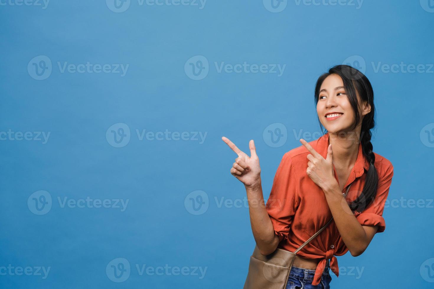 Retrato de joven asiática sonriendo con expresión alegre, muestra algo sorprendente en el espacio en blanco en ropa casual y de pie aislado sobre fondo azul. concepto de expresión facial. foto