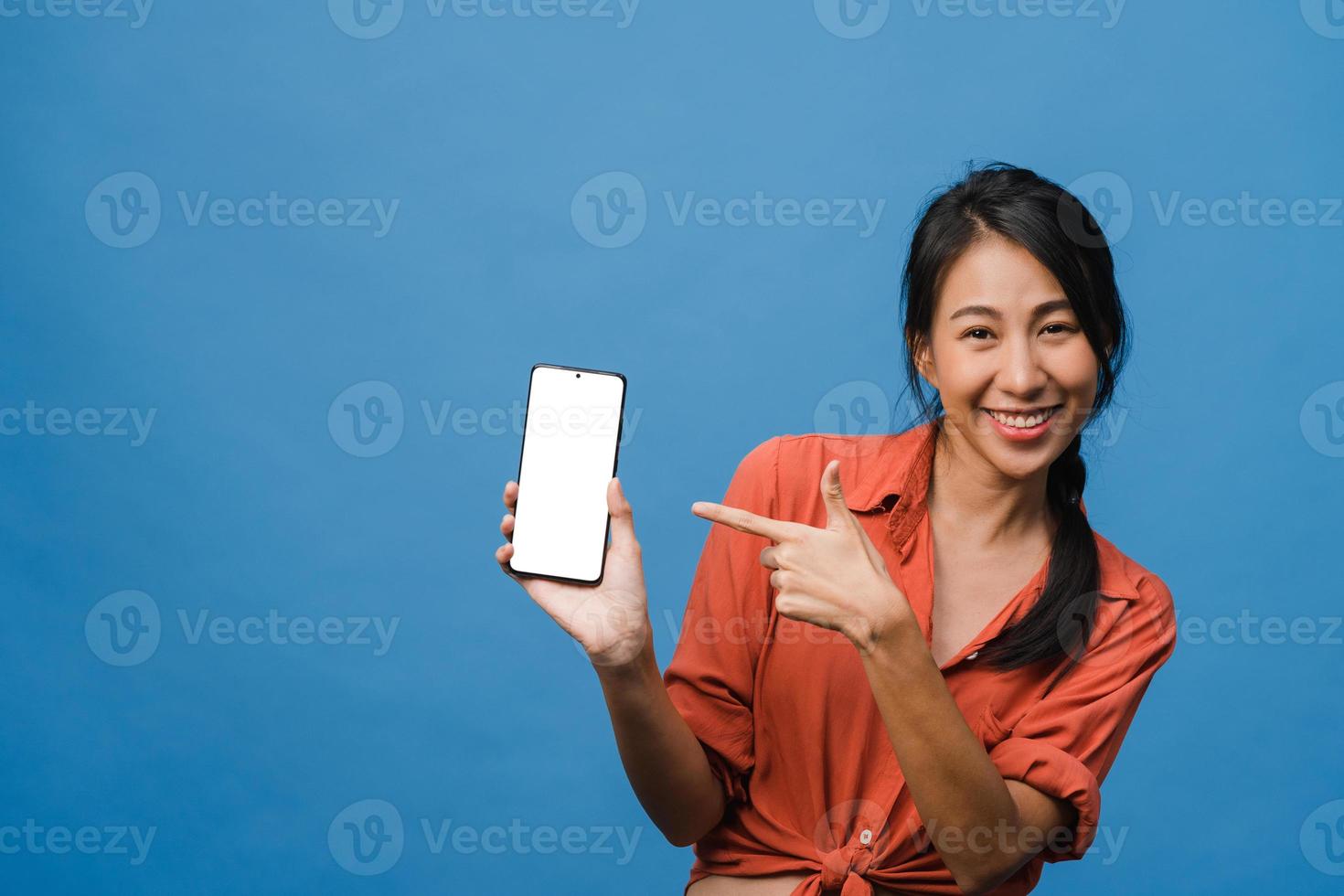 Joven asiática muestra la pantalla vacía del teléfono inteligente con expresión positiva, sonríe ampliamente, vestida con ropa casual sintiendo felicidad sobre fondo azul. teléfono móvil con pantalla en blanco en mano femenina. foto