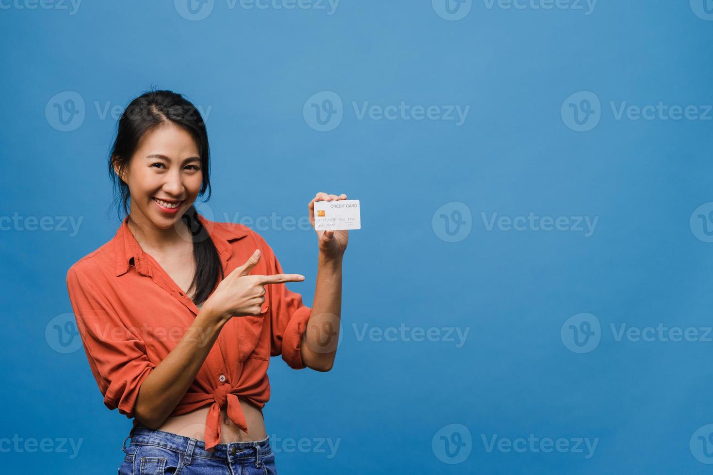 Joven mujer asiática muestra una tarjeta bancaria de crédito con expresión positiva, sonríe ampliamente, vestida con ropa informal sintiendo felicidad y parada aislada sobre fondo azul. concepto de expresión facial. foto