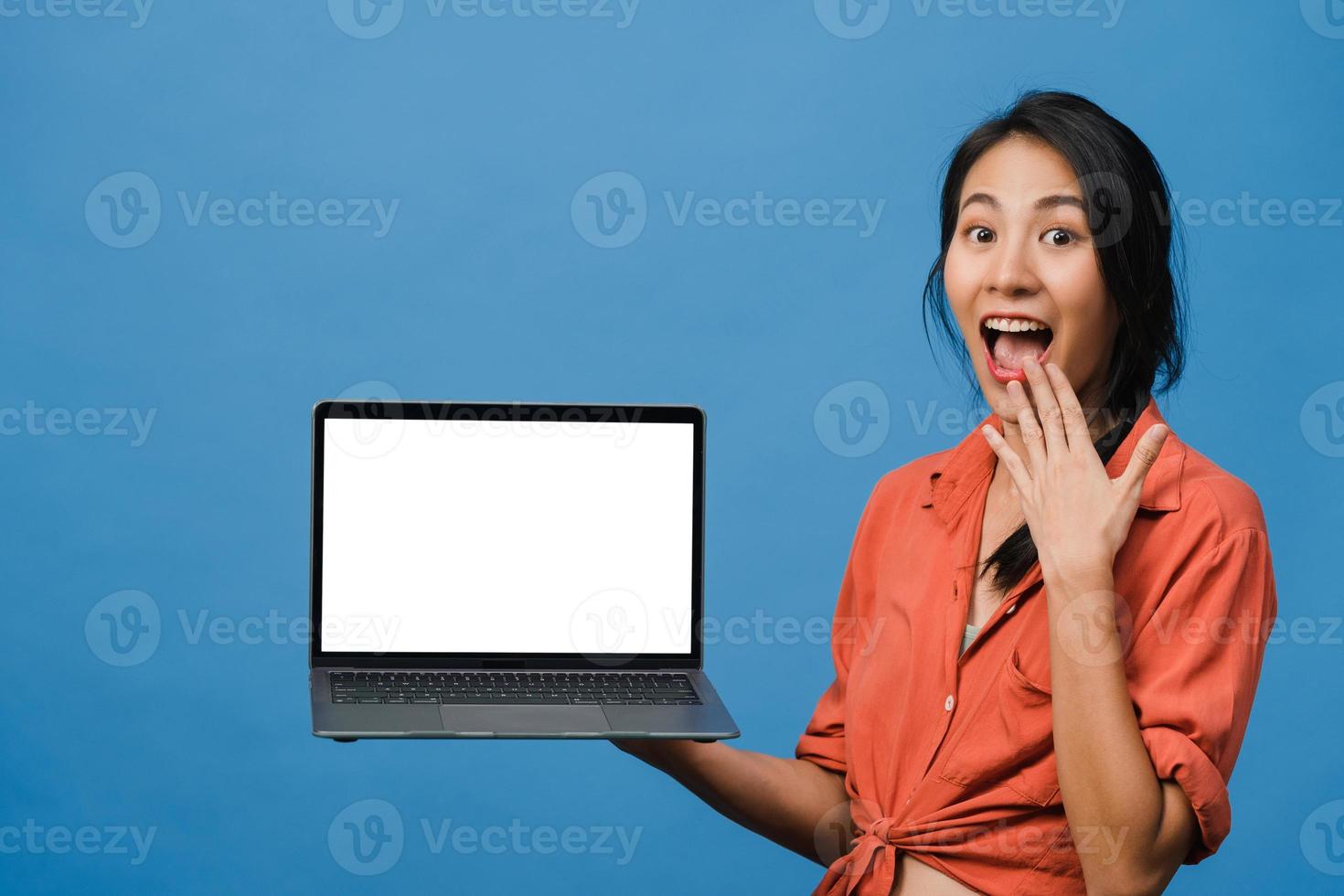 Young Asia lady show empty laptop screen with positive expression, smiles broadly, dressed in casual clothing feeling happiness isolated on blue background. Computer with white screen in female hand. photo