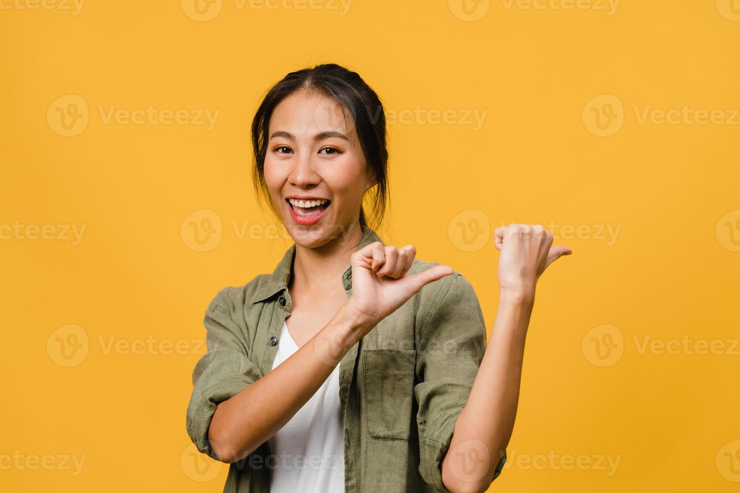 Portrait of young Asian lady smiling with cheerful expression, shows something amazing at blank space in casual cloth and looking at camera isolated over yellow background. Facial expression concept. photo