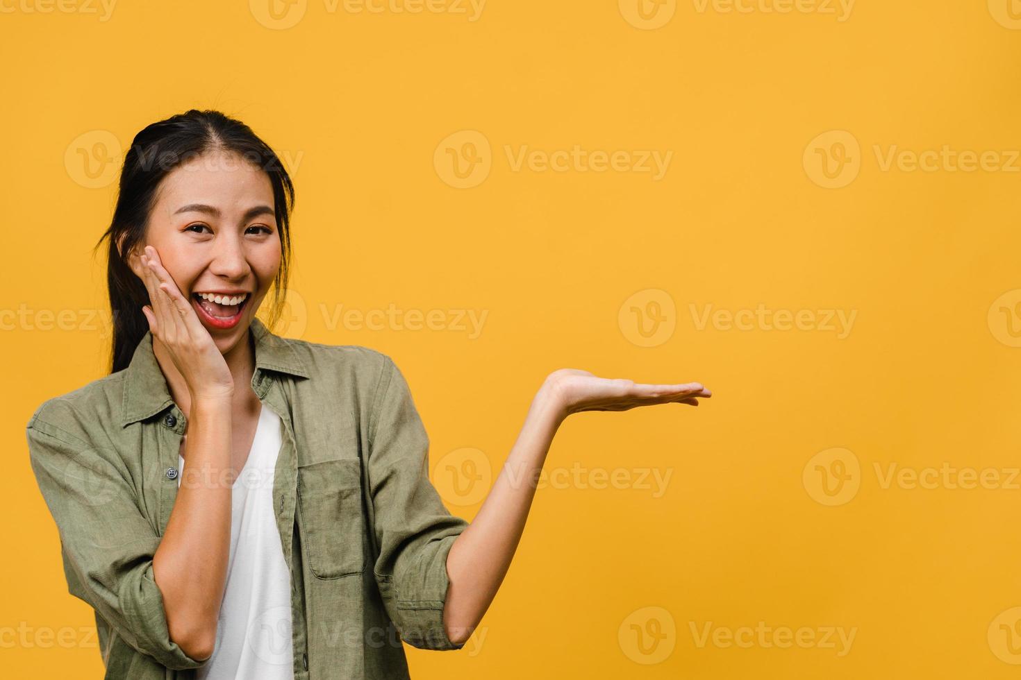 Retrato de joven asiática sonriendo con expresión alegre, muestra algo sorprendente en el espacio en blanco en un paño casual y mirando a cámara aislada sobre fondo amarillo. concepto de expresión facial. foto