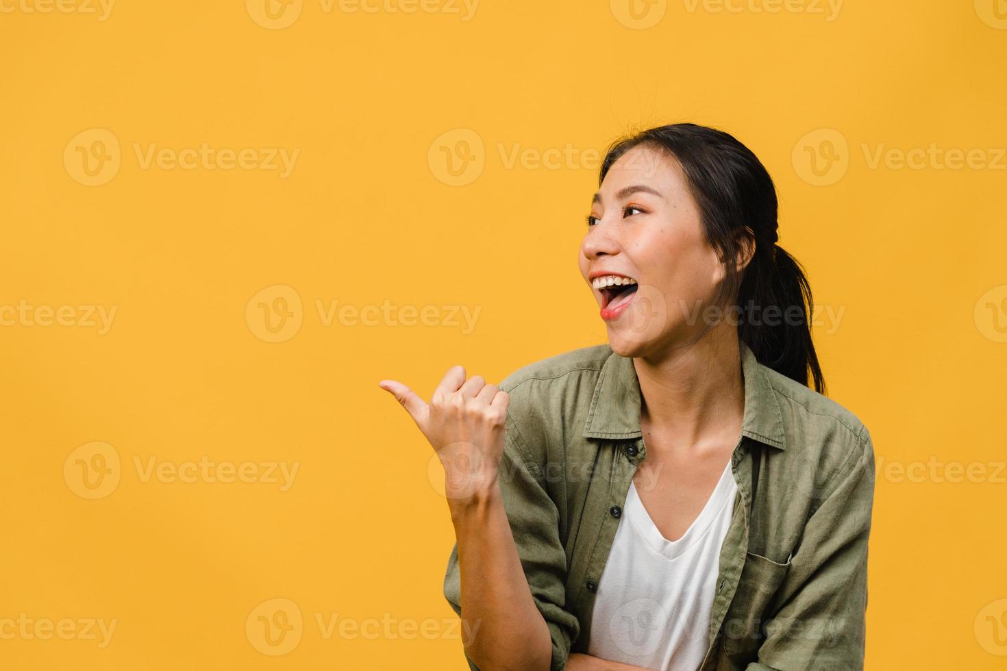 Portrait of young Asian lady smiling with cheerful expression, shows something amazing at blank space in casual clothing and standing isolated over yellow background. Facial expression concept. photo