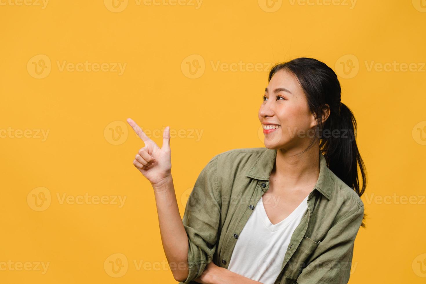 Portrait of young Asian lady smiling with cheerful expression, shows something amazing at blank space in casual clothing and standing isolated over yellow background. Facial expression concept. photo