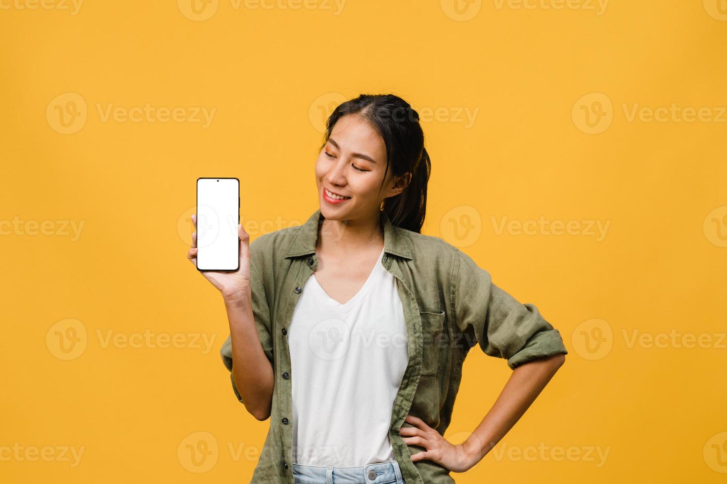 Joven asiática muestra la pantalla vacía del teléfono inteligente con expresión positiva, sonríe ampliamente, vestida con ropa casual sintiendo felicidad sobre fondo amarillo. teléfono móvil con pantalla en blanco en mano femenina. foto