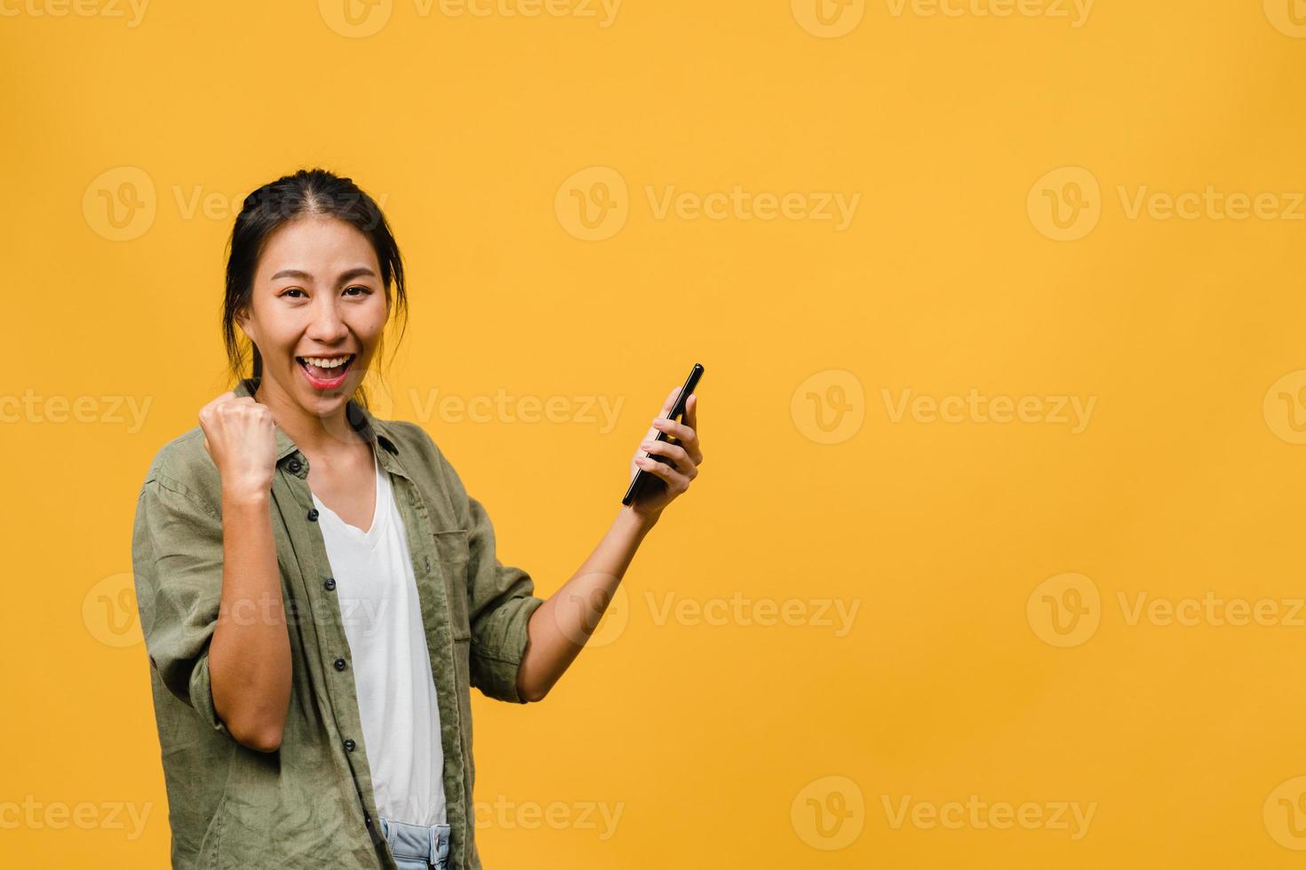 Surprised young Asia lady using mobile phone with positive expression, smiles broadly, dressed in casual clothing and stand isolated on yellow background. Happy adorable glad woman rejoices success. photo