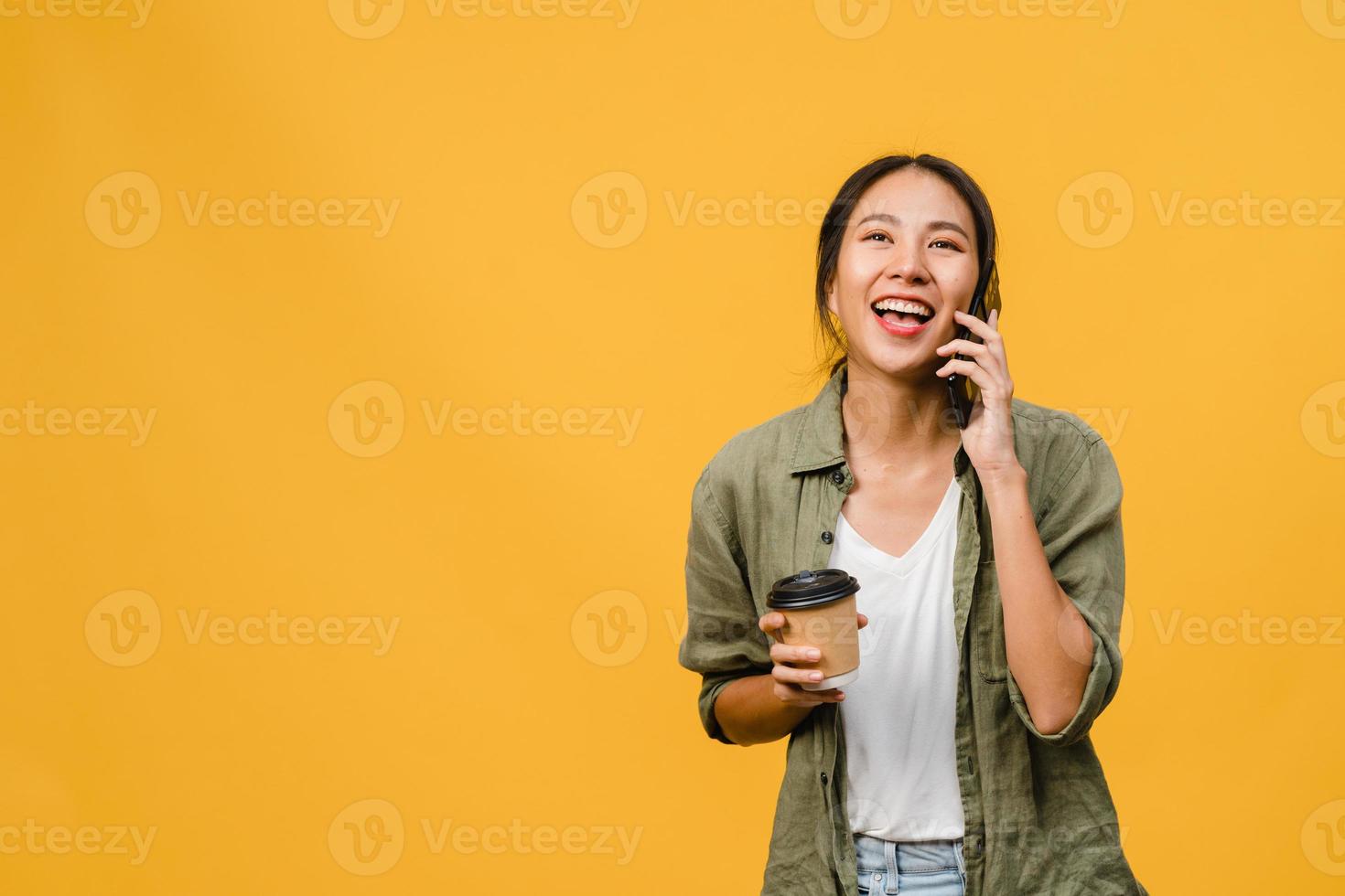 La joven asiática habla por teléfono y sostiene la taza de café con expresión positiva, sonríe ampliamente, vestida con un paño informal sintiendo felicidad y parada aislada sobre fondo amarillo. concepto de expresión facial. foto