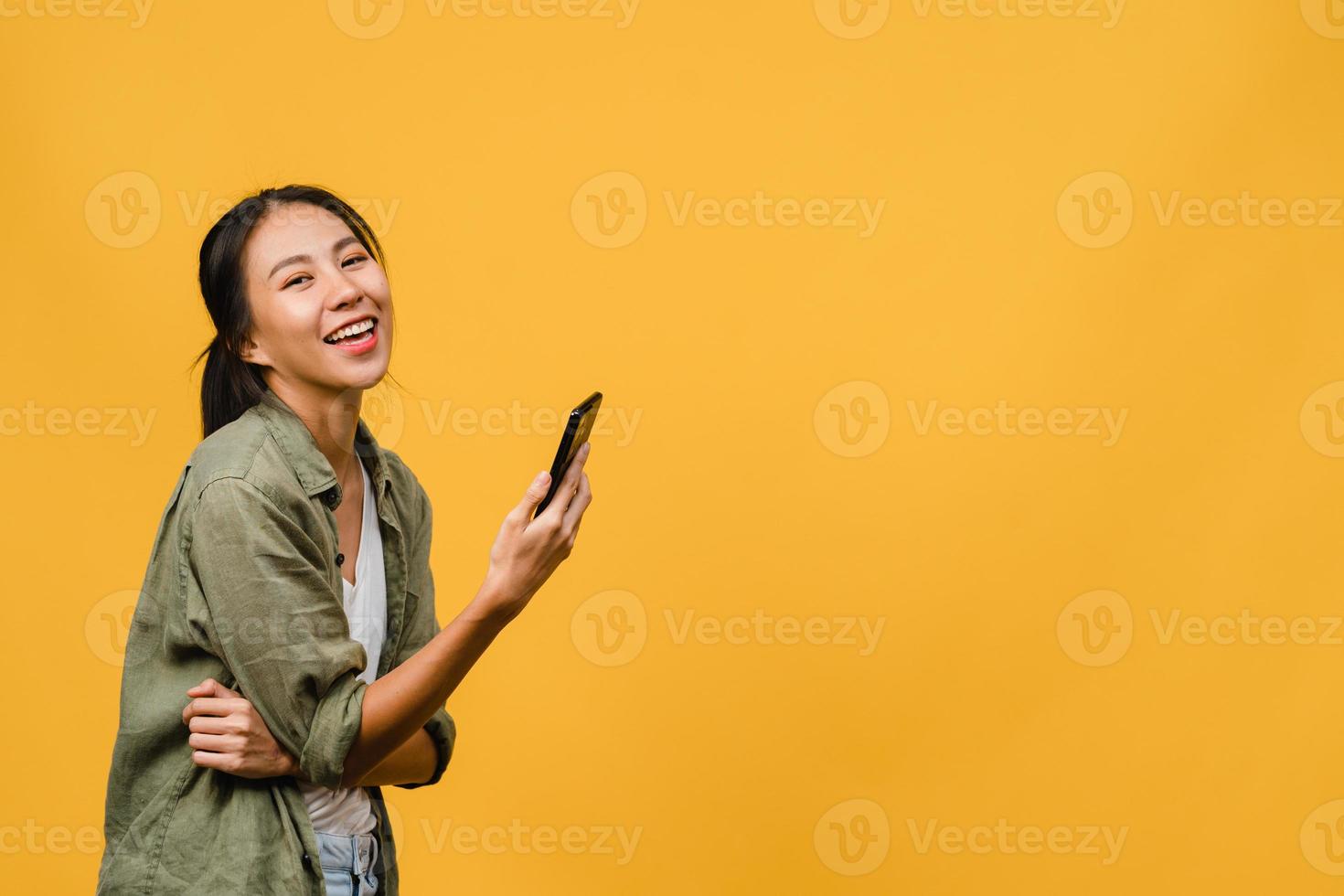 Surprised young Asia lady using mobile phone with positive expression, smile broadly, dressed in casual clothing and looking at camera on yellow background. Happy adorable glad woman rejoices success. photo