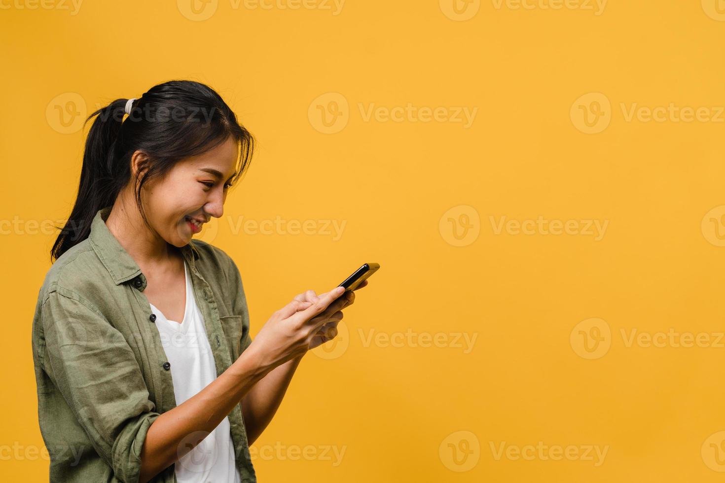 Jovencita asiática que usa el teléfono con expresión positiva, sonríe ampliamente, vestida con ropa casual sintiendo felicidad y parada aislada sobre fondo amarillo. feliz adorable mujer alegre se regocija con el éxito. foto