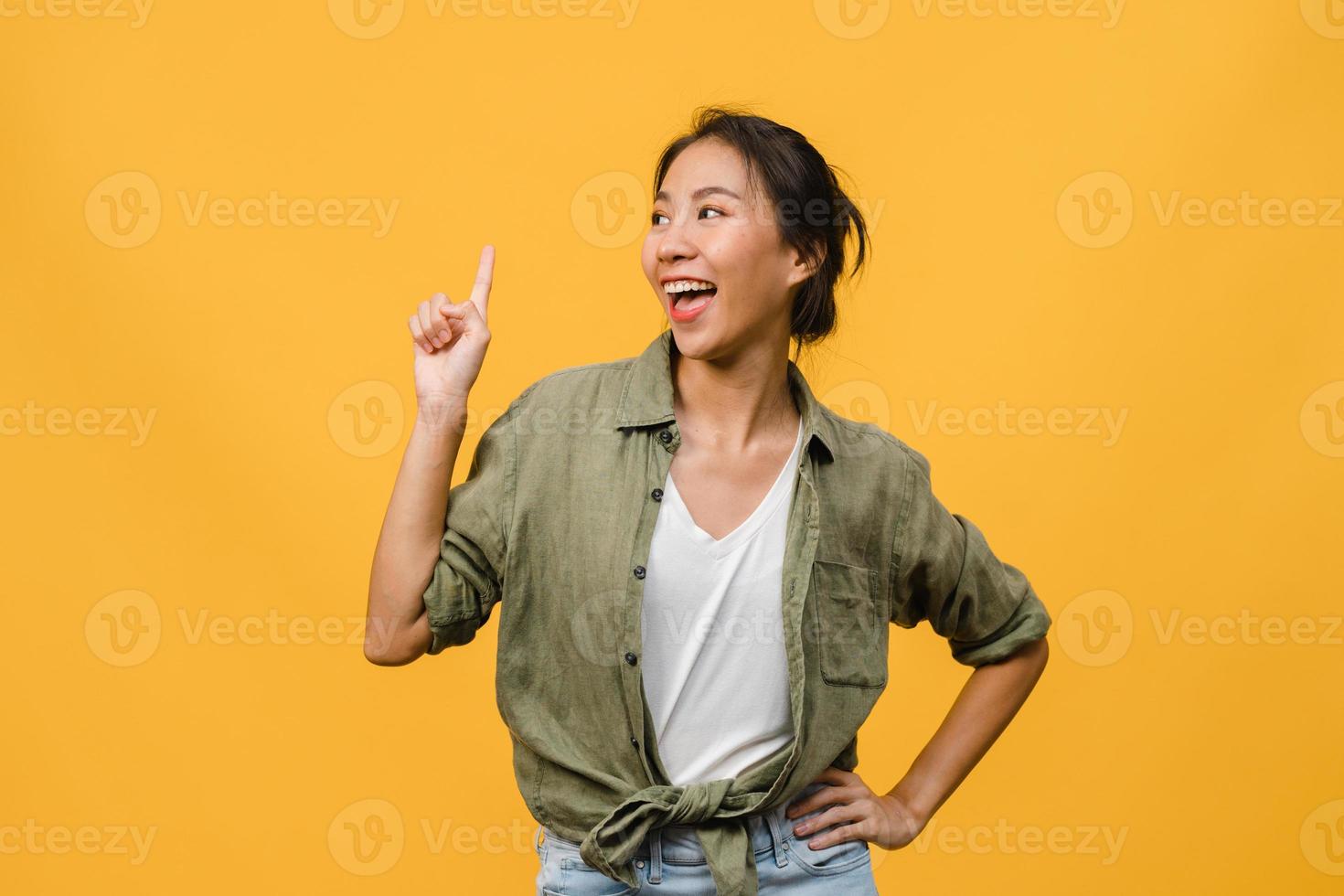 Portrait of young Asian lady smiling with cheerful expression, shows something amazing at blank space in casual clothing and standing isolated over yellow background. Facial expression concept. photo