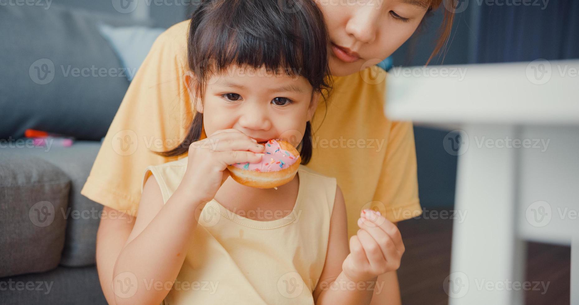 Happy cheerful Asia family mom and toddler girl eating donuts and having fun relax enjoy on couch in living room at house. Spending time together, Social distance, Quarantine for coronavirus. photo