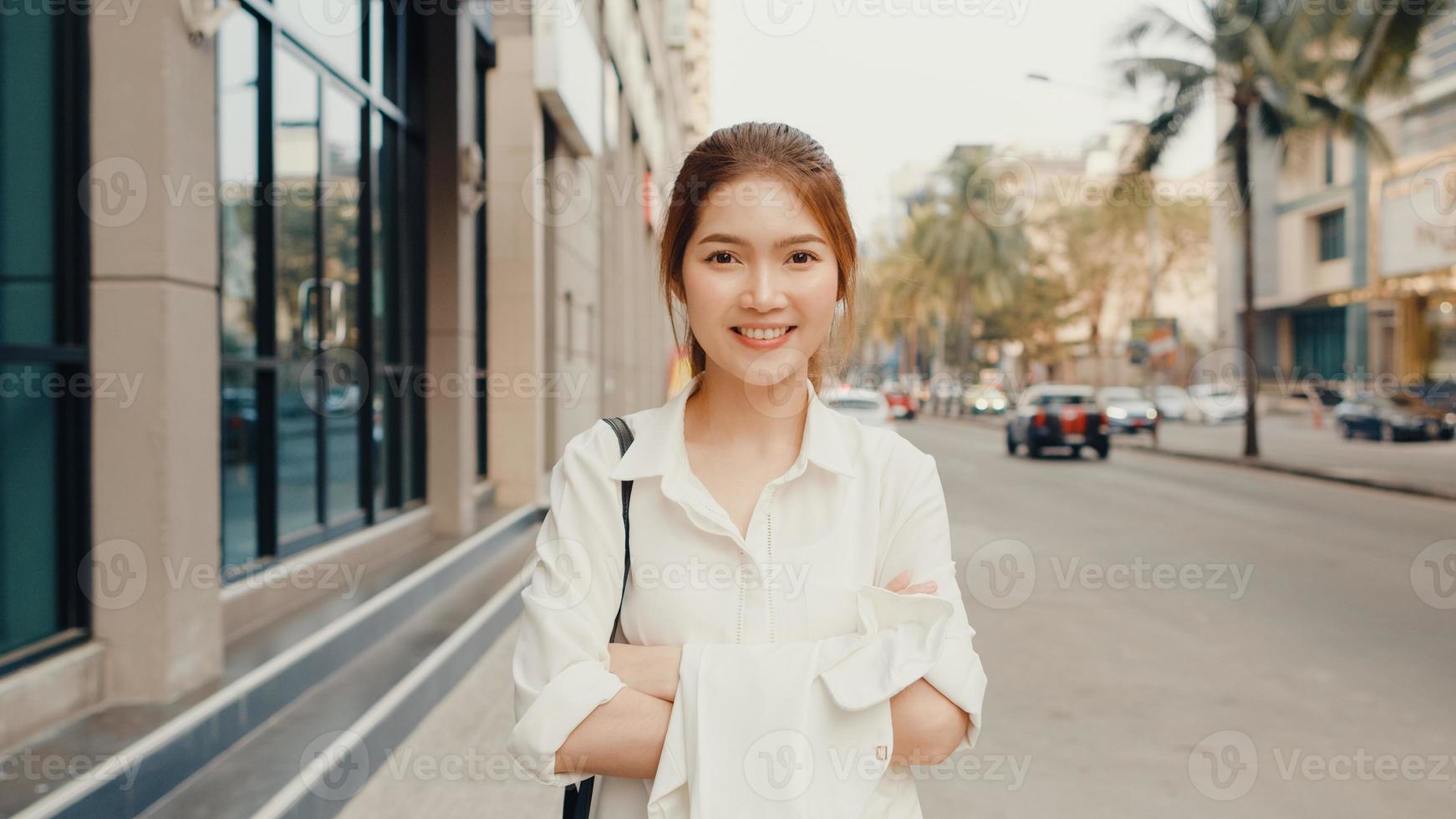 Successful young Asia businesswoman in fashion office clothes smiling and looking at camera while happy standing alone outdoors in urban modern city in the morning. Business on the go concept. photo