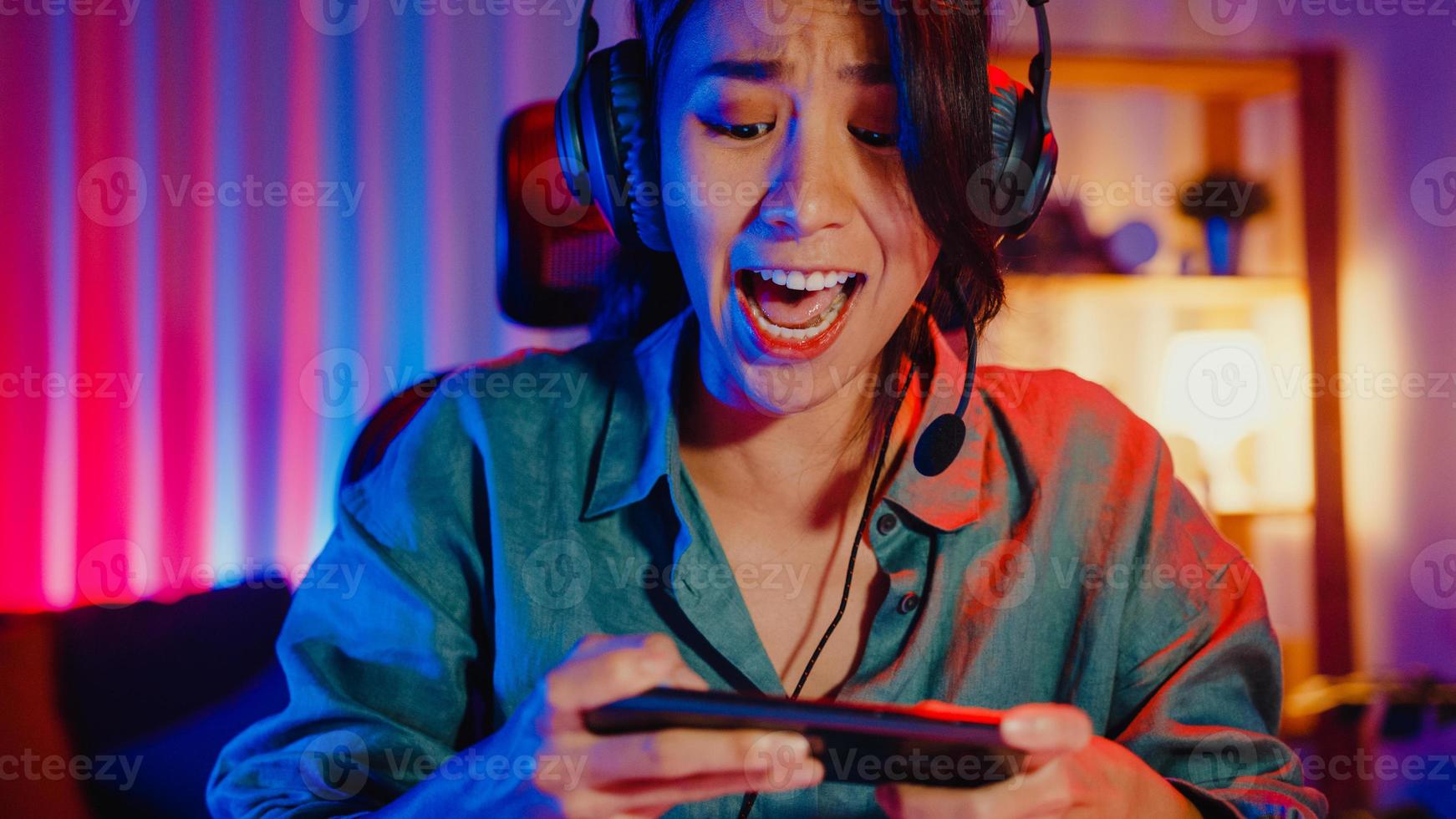 Happy asia girl gamer wear headphone competition play video game online with smartphone colorful neon lights in living room at home. Esport streaming game online, Home quarantine activity concept. photo