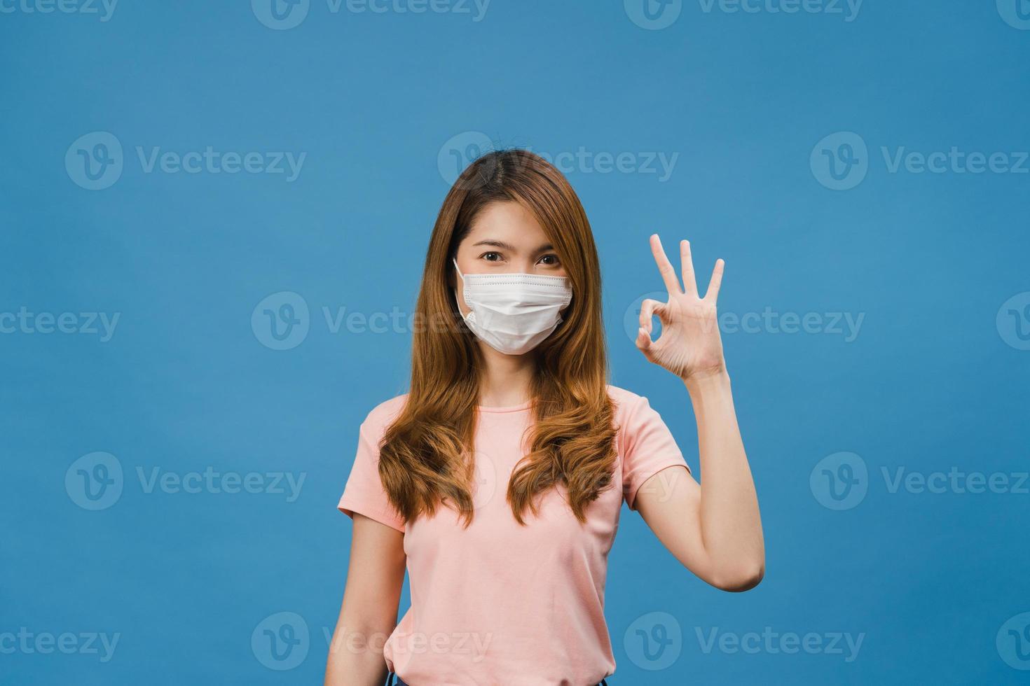 Chica joven de Asia con mascarilla médica gesticulando ok firmar vestida con ropa casual y mirar a cámara aislada sobre fondo azul. autoaislamiento, distanciamiento social, cuarentena por coronavirus. foto