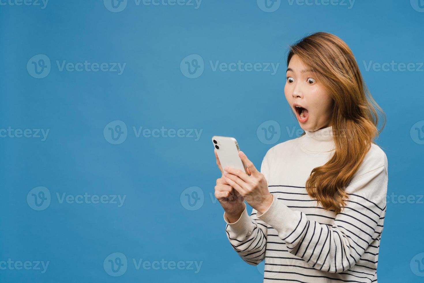 Surprised young Asia lady using mobile phone with positive expression, smiles broadly, dressed in casual clothing and standing isolated on blue background. Happy adorable glad woman rejoices success. photo