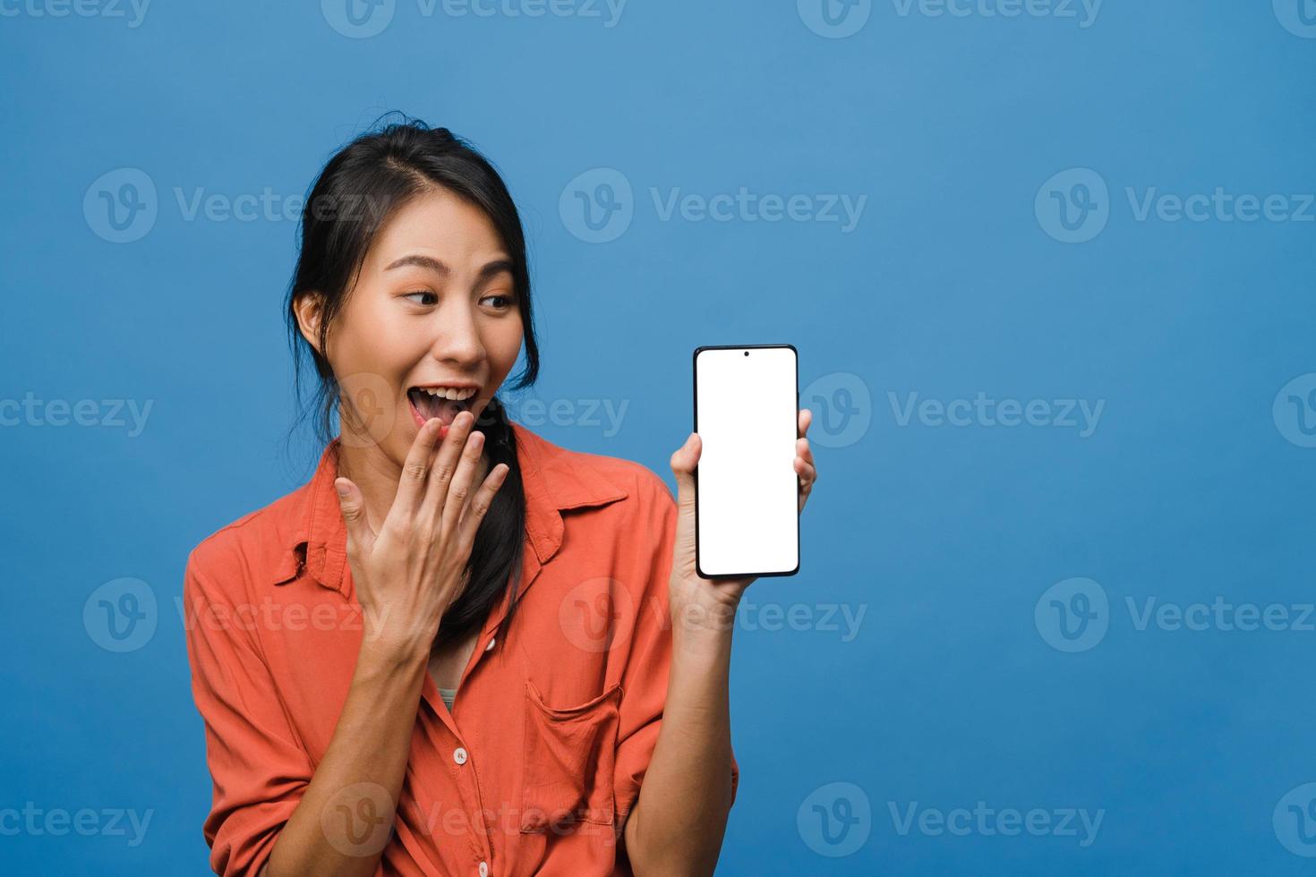 Joven asiática muestra la pantalla vacía del teléfono inteligente con expresión positiva, sonríe ampliamente, vestida con ropa casual sintiendo felicidad sobre fondo azul. teléfono móvil con pantalla en blanco en mano femenina. foto