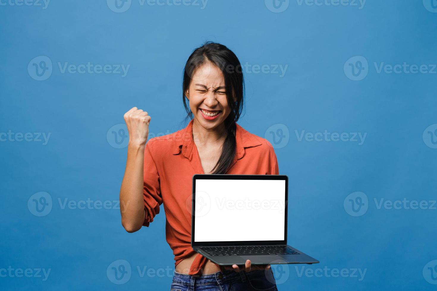 Joven asiática muestra una pantalla de computadora portátil vacía con expresión positiva, sonríe ampliamente, vestida con ropa casual sintiendo felicidad aislada sobre fondo azul. computadora con pantalla blanca en mano femenina. foto