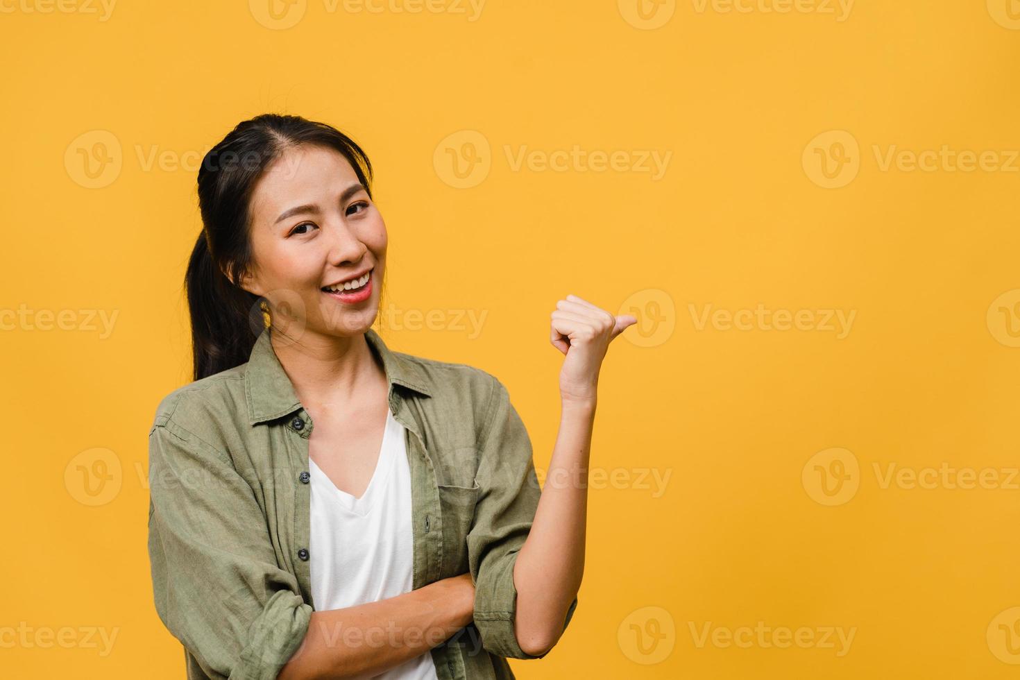 Portrait of young Asian lady smiling with cheerful expression, shows something amazing at blank space in casual cloth and looking at camera isolated over yellow background. Facial expression concept. photo