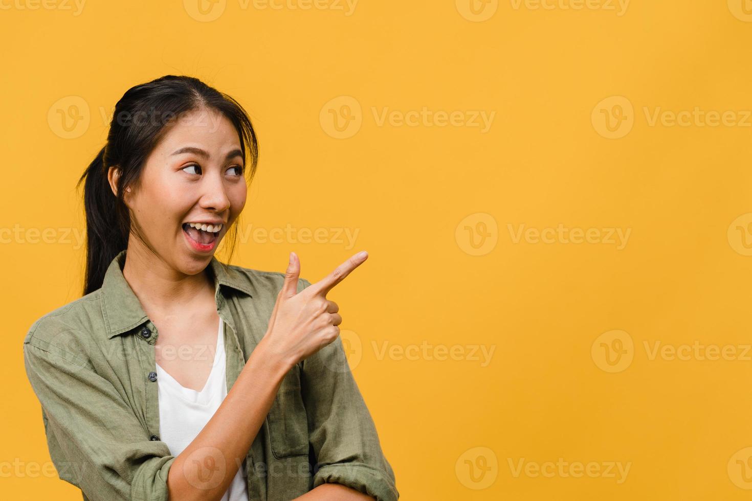 Portrait of young Asian lady smiling with cheerful expression, shows something amazing at blank space in casual clothing and standing isolated over yellow background. Facial expression concept. photo
