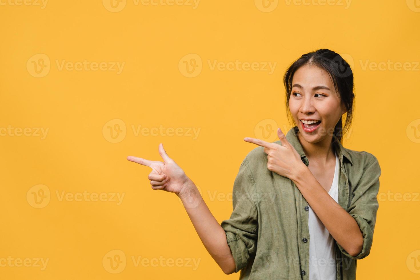 Portrait of young Asian lady smiling with cheerful expression, shows something amazing at blank space in casual clothing and standing isolated over yellow background. Facial expression concept. photo