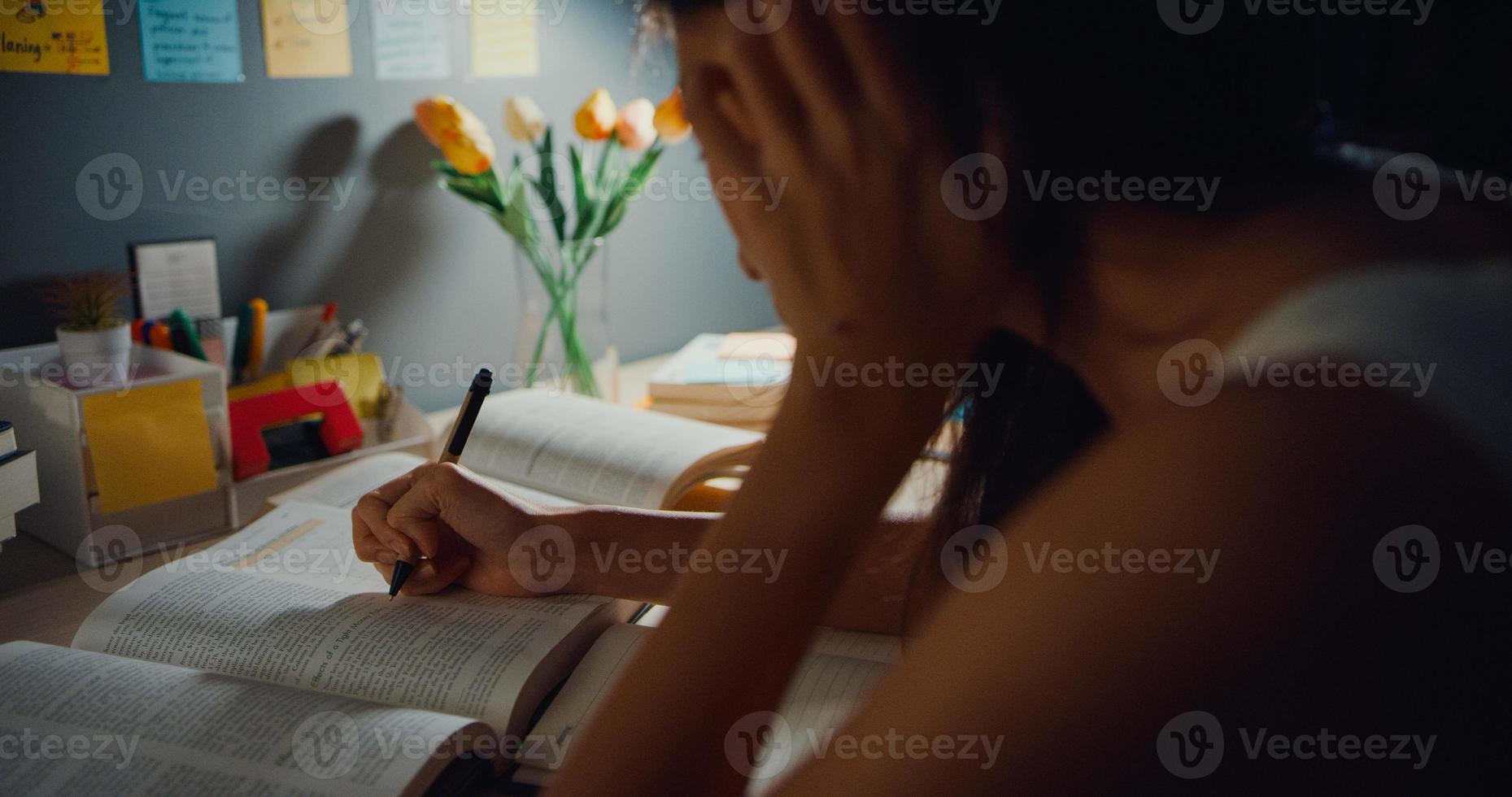 Young Asia teen girl student distance learning lesson and doing homework sitting at desk in living room at house night. Work from home, Social distancing, quarantine for corona virus prevention. photo