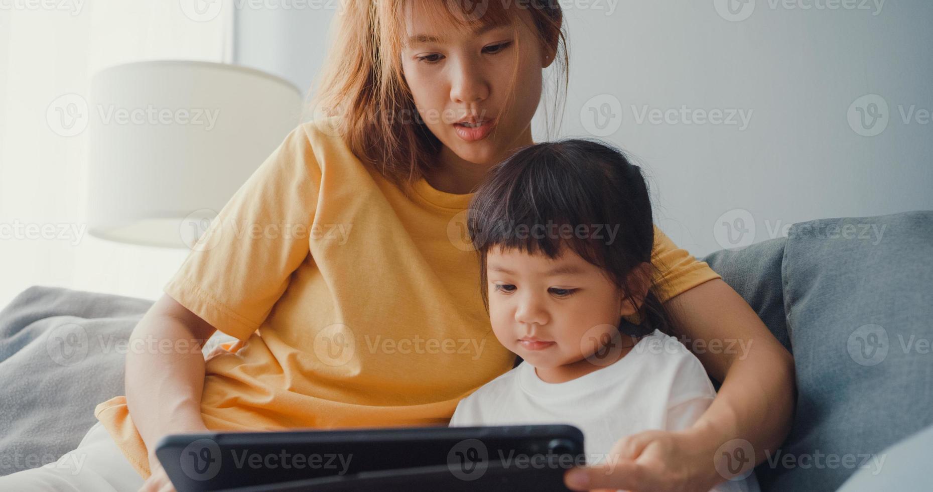 Feliz madre de familia asiática alegre y niño lindo usando dibujos animados de interés de tableta digital y ver películas divirtiéndose relajarse en el sofá en la sala de estar de la casa. pasar tiempo juntos, cuarentena por coronavirus. foto