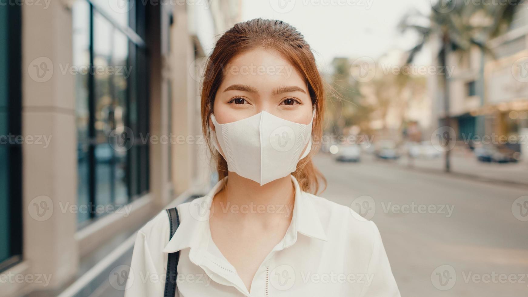 Exitosa empresaria asiática joven en ropa de oficina de moda usa mascarilla médica sonriendo y mirando a la cámara mientras está feliz de pie sola al aire libre en la ciudad moderna urbana. concepto de negocio en movimiento. foto