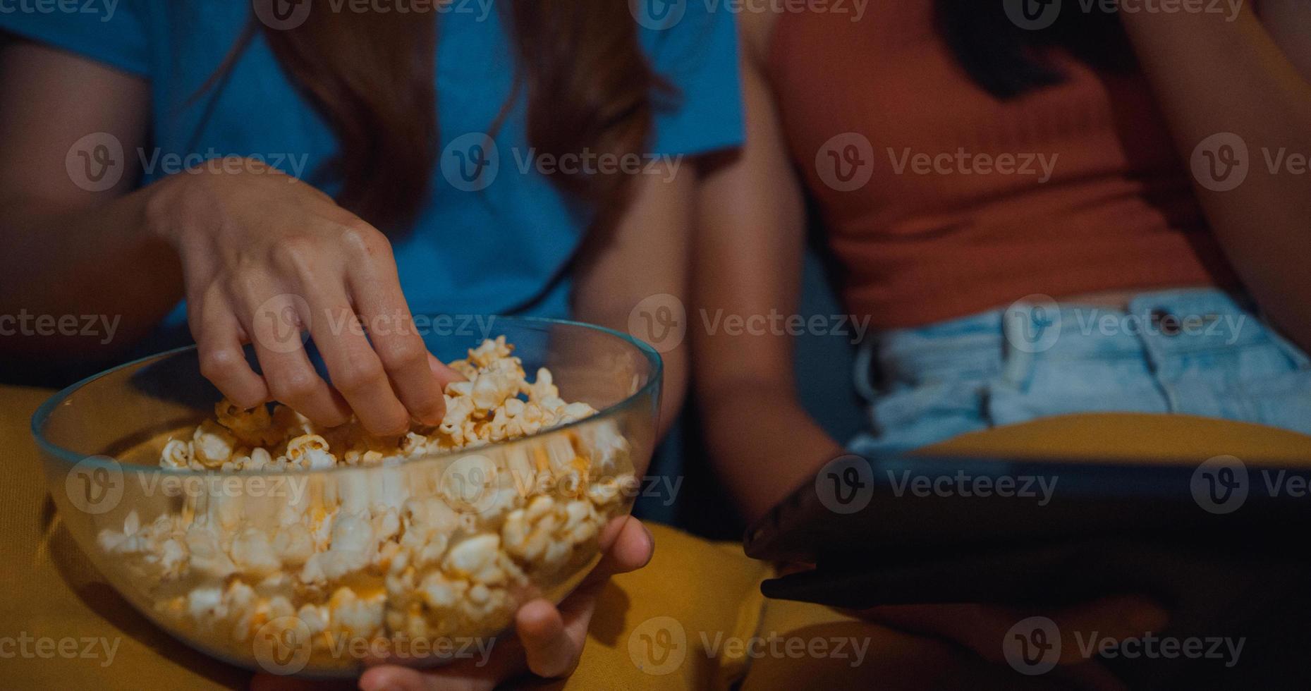 atractivas damas asiáticas con casual disfrutan de un momento feliz, miran entretenimiento de películas en línea en una tableta, comen un sitio de palomitas de maíz en el sofá, sala de estar en casa por la noche. concepto de cuarentena de actividad de estilo de vida. foto