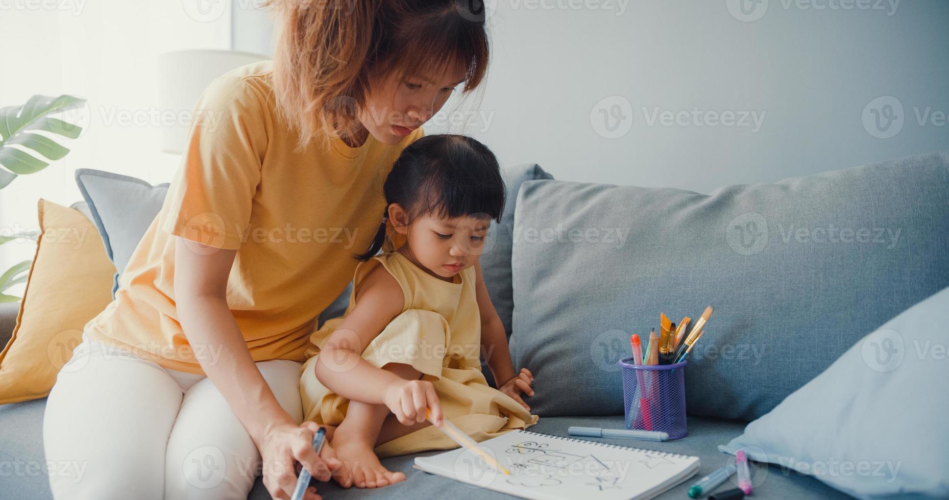 feliz, alegre, asia, familia, mamá, enseñar, niña, pintura, uso, álbum, y, colorido, lápices, divirtiéndose, relajarse, en, sofá, en, sala, en, casa. pasar tiempo juntos, distancia social, cuarentena por coronavirus. foto