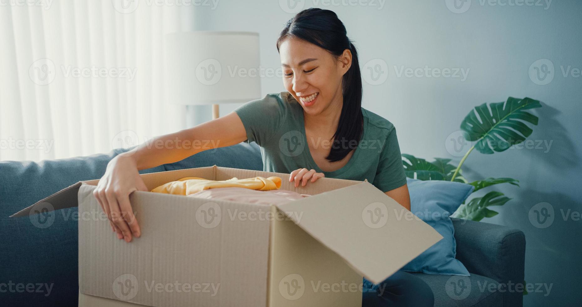 Happy asia lady open cardboard box package exciting and enjoy trying and matching with quality of fashion cloth product from online market in living room at home. Online shopping and delivery concept. photo