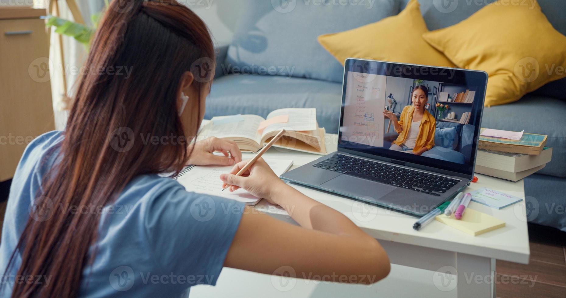 joven asiática con videollamada de computadora portátil de uso casual aprender en línea con el maestro escribir conferencia cuaderno sala de estar en casa. Aislar el concepto de pandemia de coronavirus de educación en línea e-learning. foto