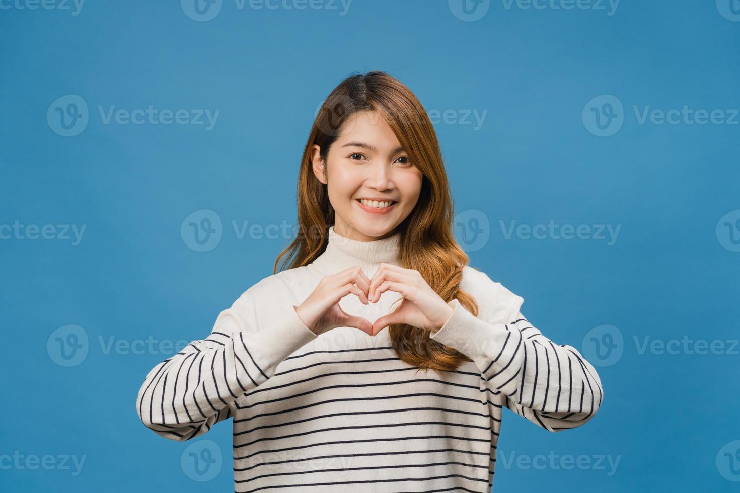 Young Asia lady with positive expression, shows hands gesture in heart shape, dressed in casual clothing and looking at camera isolated on blue background. Happy adorable glad woman rejoices success. photo