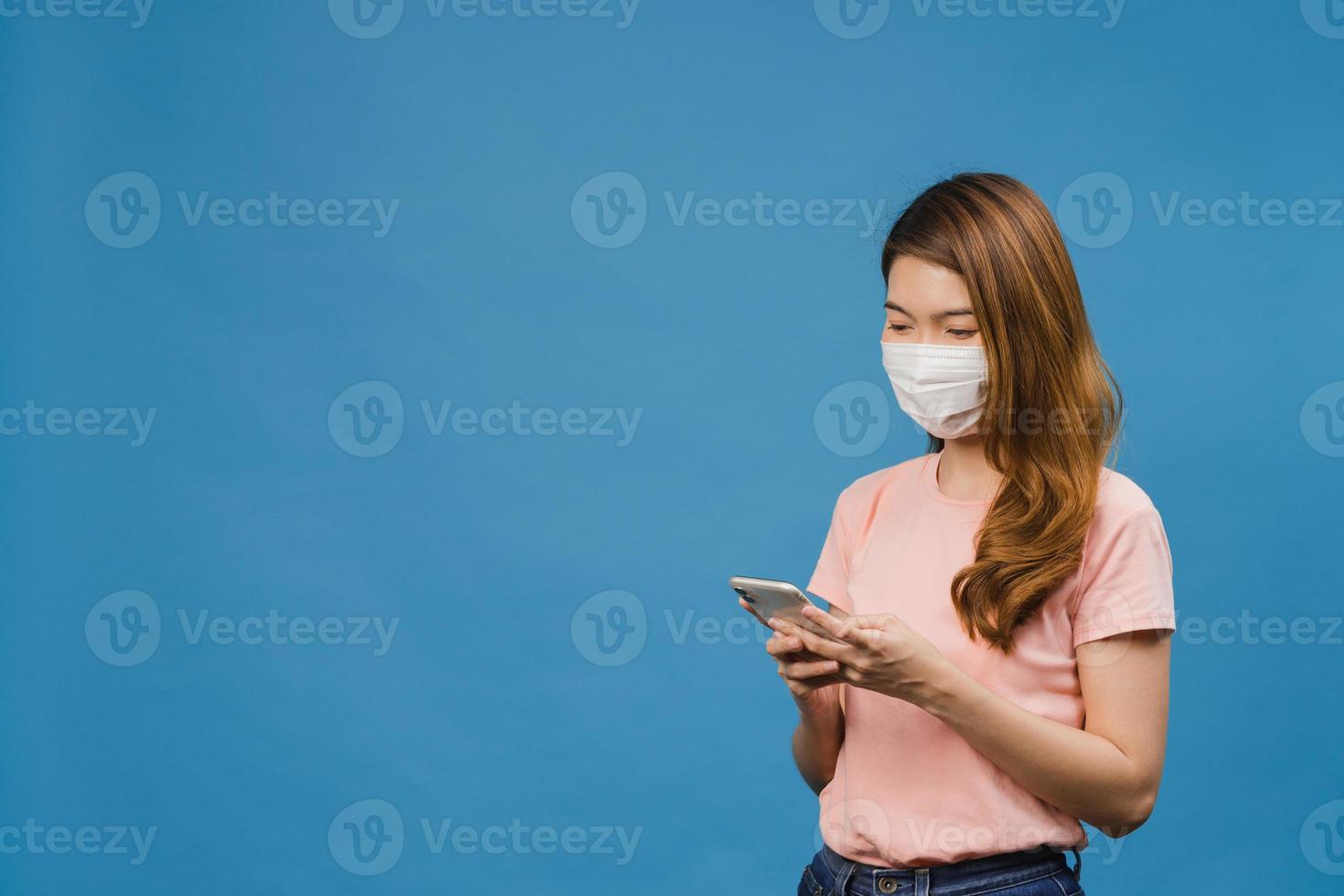 Young Asia girl wearing medical face mask using mobile phone with dressed in casual clothing isolated on blue background. Self-isolation, social distancing, quarantine for corona virus prevention. photo